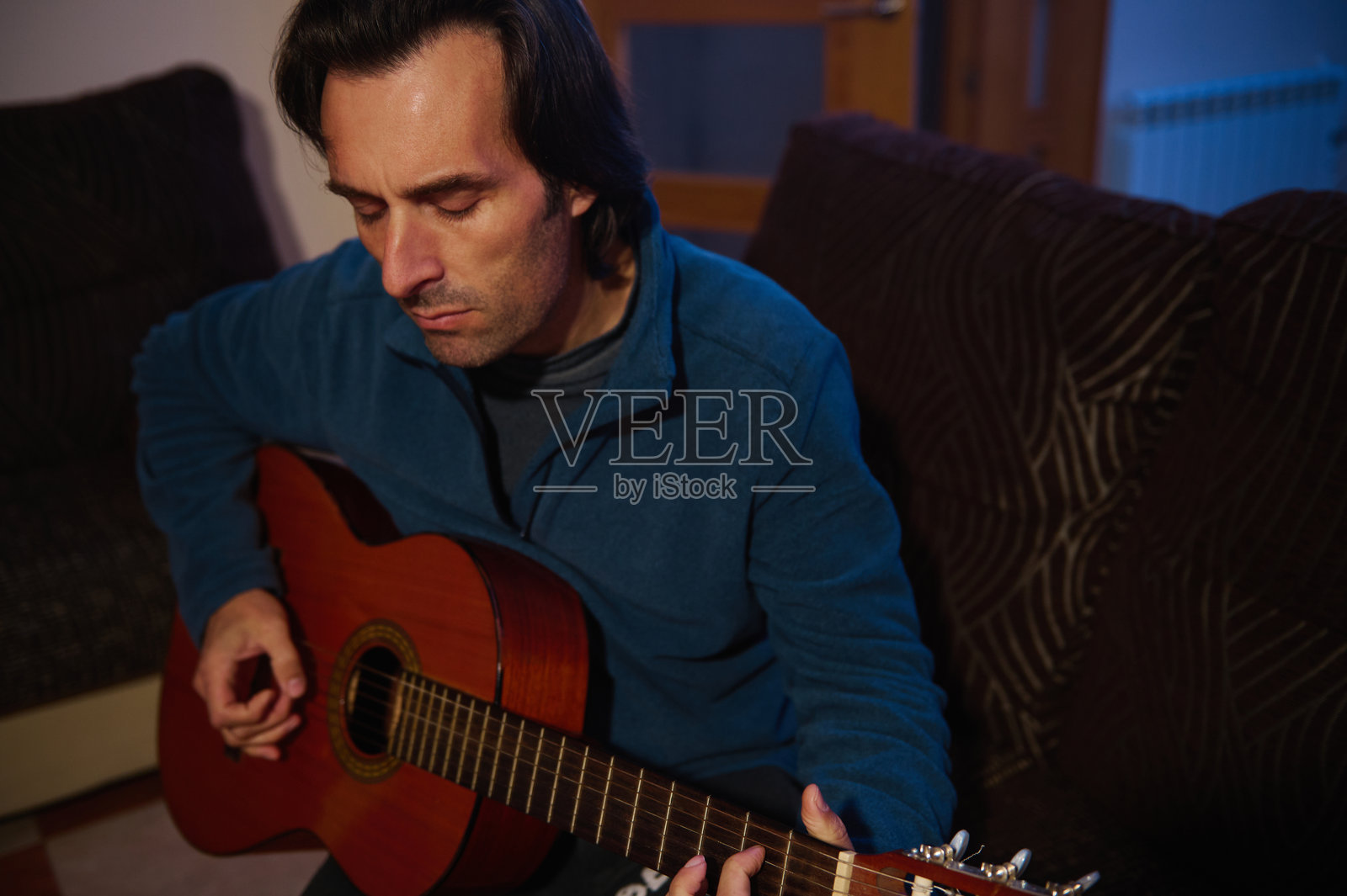 一个男人在舒适的客厅环境中专注地弹奏着原声吉他，穿着蓝色毛衣。捕捉音乐与放松在温馨家居环境中的本质。照片摄影图片