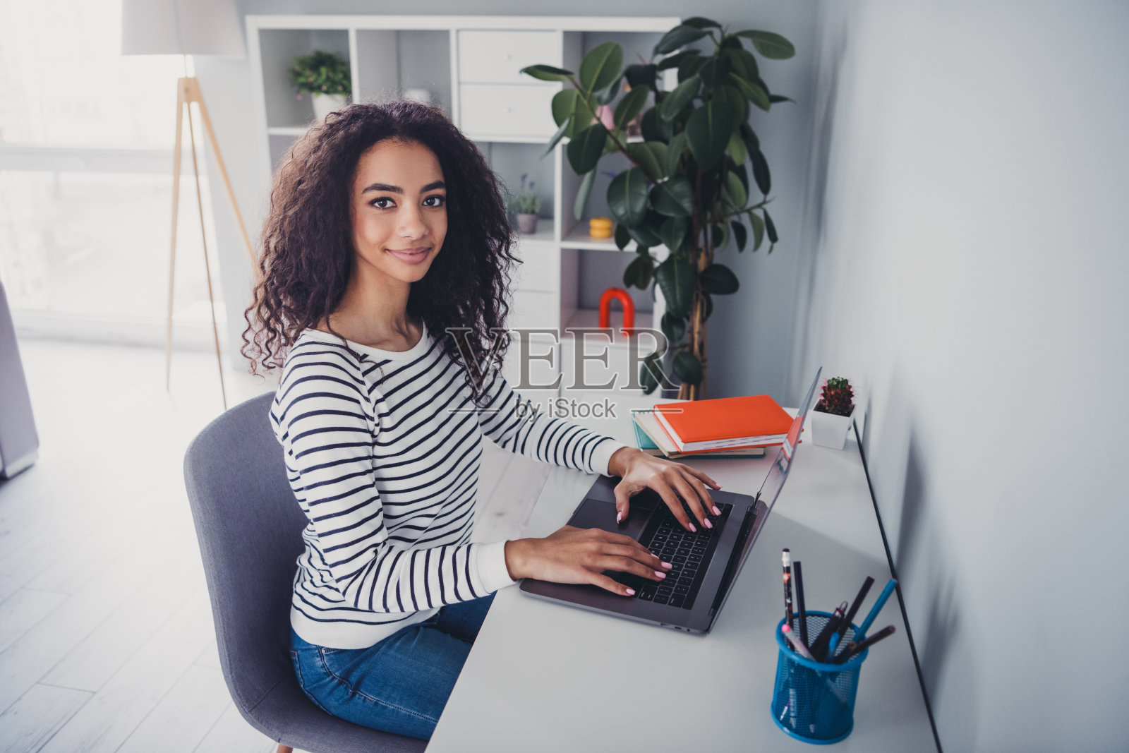 年轻女孩坐在椅子上，使用笔记本电脑，穿着条纹毛衣，居住在公寓的室内客厅。照片摄影图片