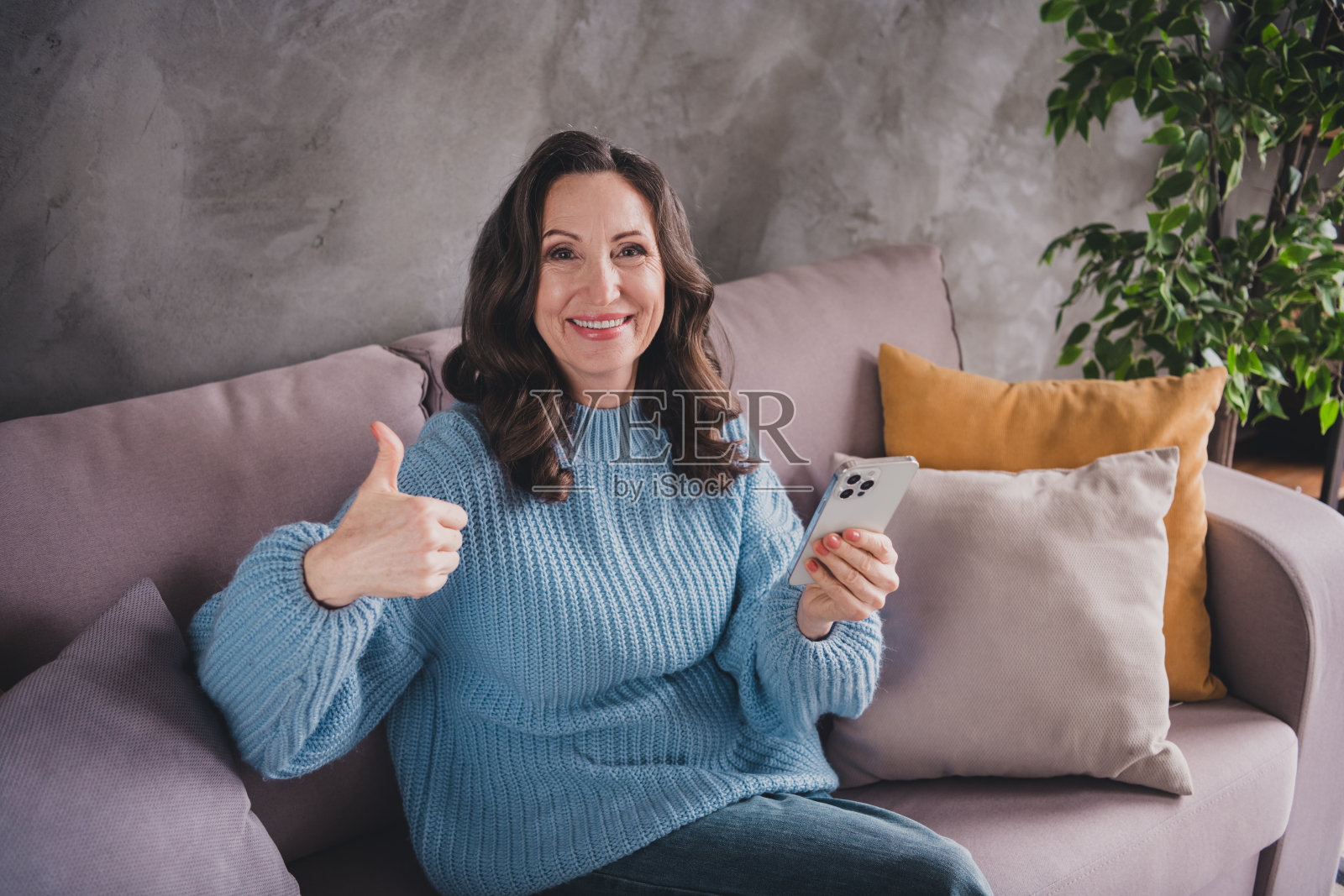 一位留着棕色头发、穿着蓝色毛衣的老年女性在室内使用智能手机，竖起大拇指。照片摄影图片