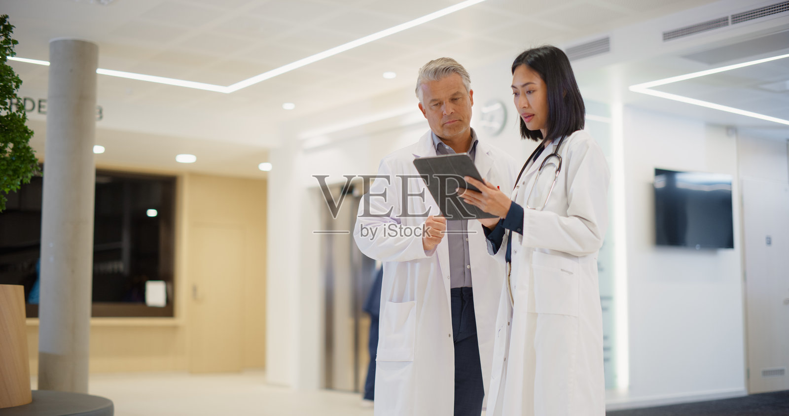 白人医生与年轻的亚洲女性医生在现代医院大堂讨论平板电脑上的病人数据，进行轻松的医疗对话并讨论在线医疗数据。照片摄影图片