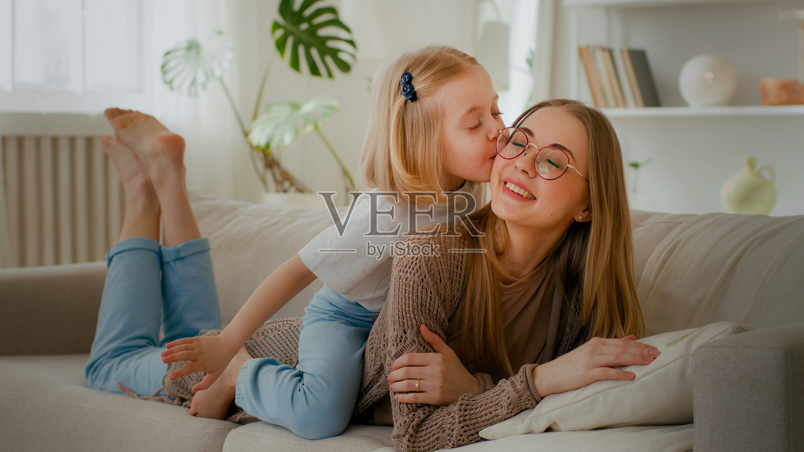 快乐的母亲与女儿亲密无间，白人母亲躺在沙发上，女儿骑在背上，亲吻妈妈，温馨的交流与甜蜜的亲情。照片摄影图片