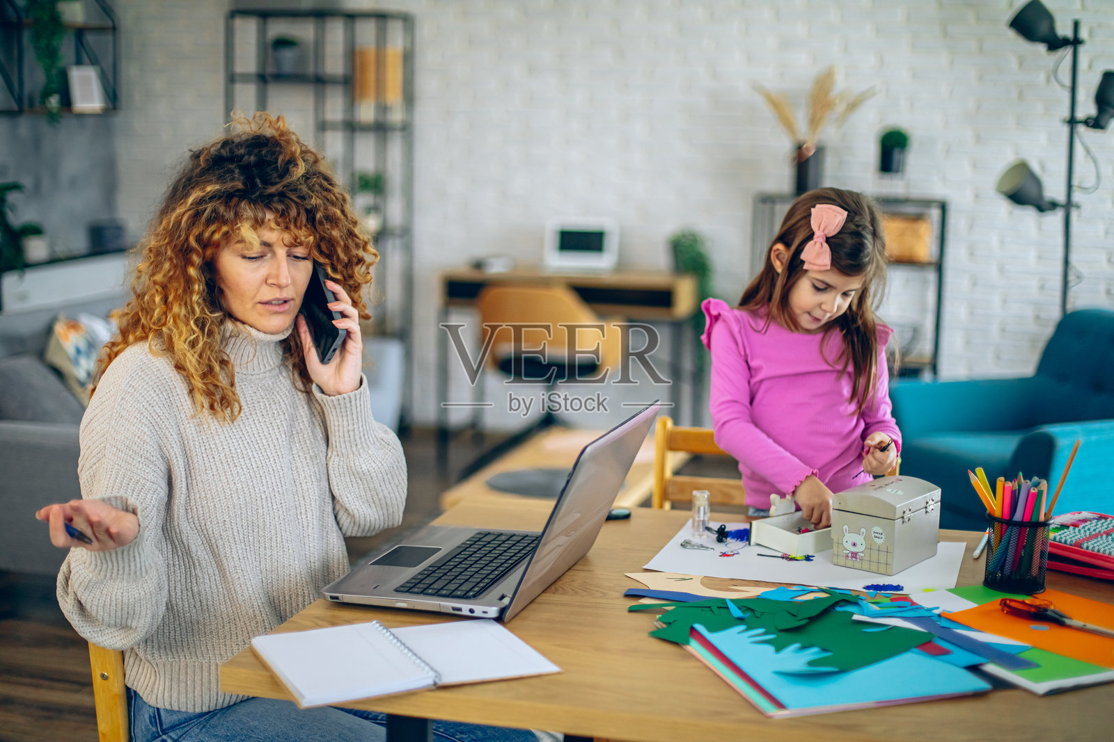 母亲在餐桌上使用笔记本电脑和手机，而女儿则在玩五颜六色的纸张，展示了家庭办公室中的工作与生活平衡。照片摄影图片