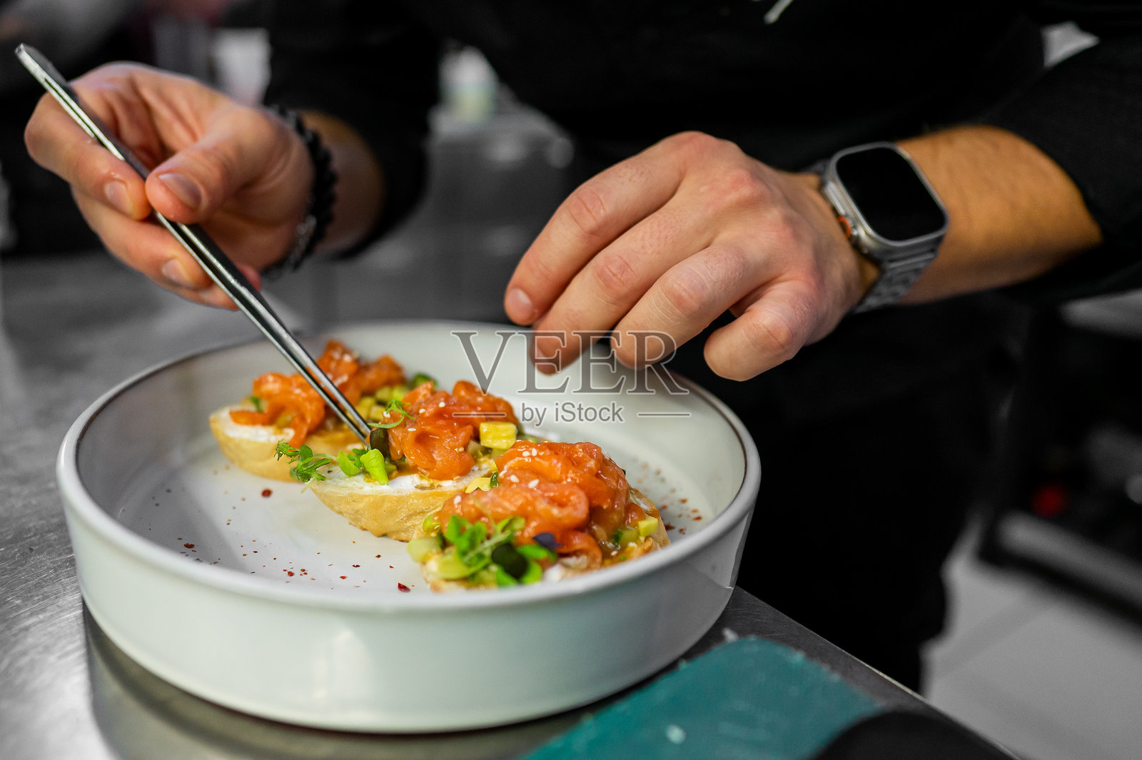 一位厨师在白色盘子上精心装饰美味的虾开胃小菜，展示了他在充满活力的厨房环境中的烹饪技巧。非常适合与美食和烹饪相关的主题。照片摄影图片