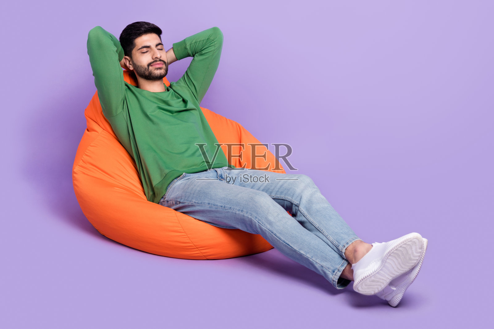 年轻的拉丁裔男子穿着时尚的绿色服装，躺在豆袋椅上，悠闲地做白日梦，背景为紫色。照片摄影图片