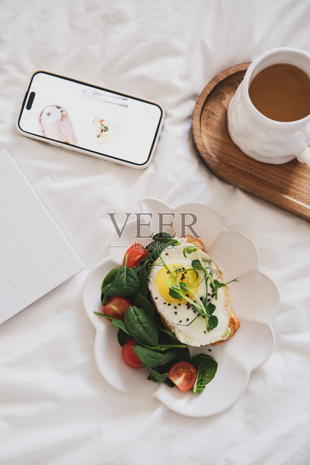 一顿丰盛的早餐，包括美味的鳄梨吐司配鸡蛋和菠菜、番茄、香气四溢的茶，以及放在舒适白色床上的智能手机。照片摄影图片