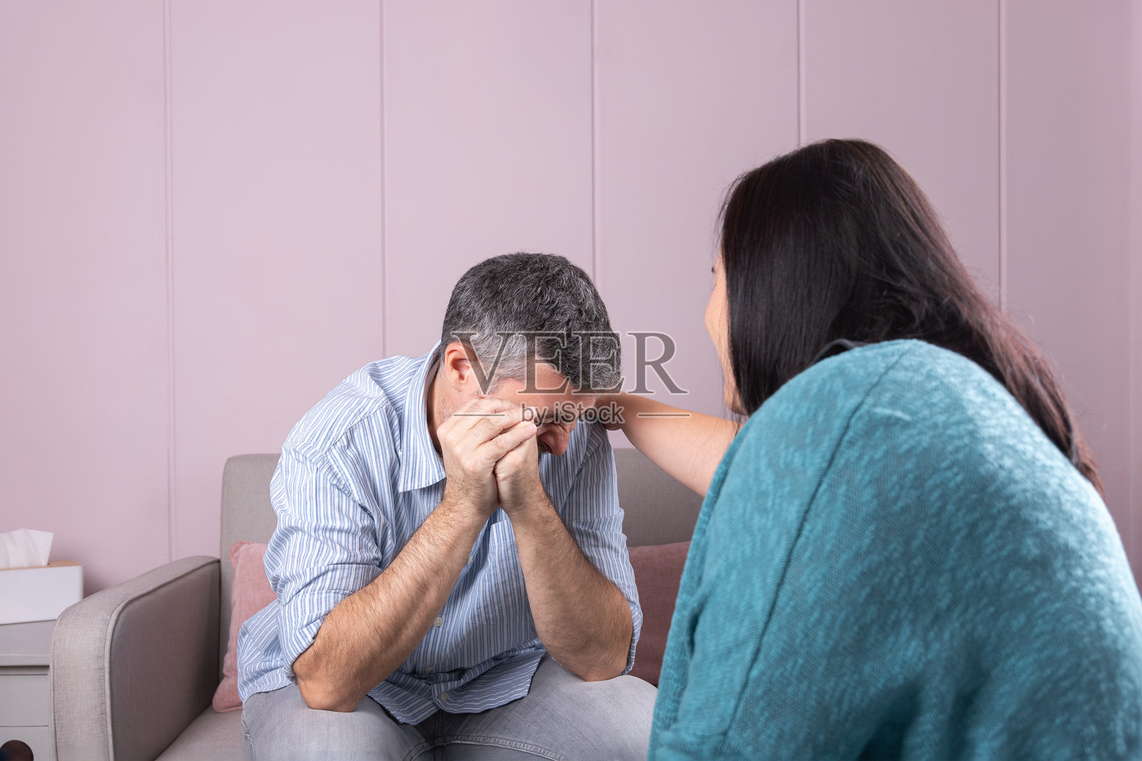 一名男子在心理咨询中哭泣，双手捂住脸，心理治疗师背对着他，在温暖而专业的环境中陪伴着他。照片摄影图片