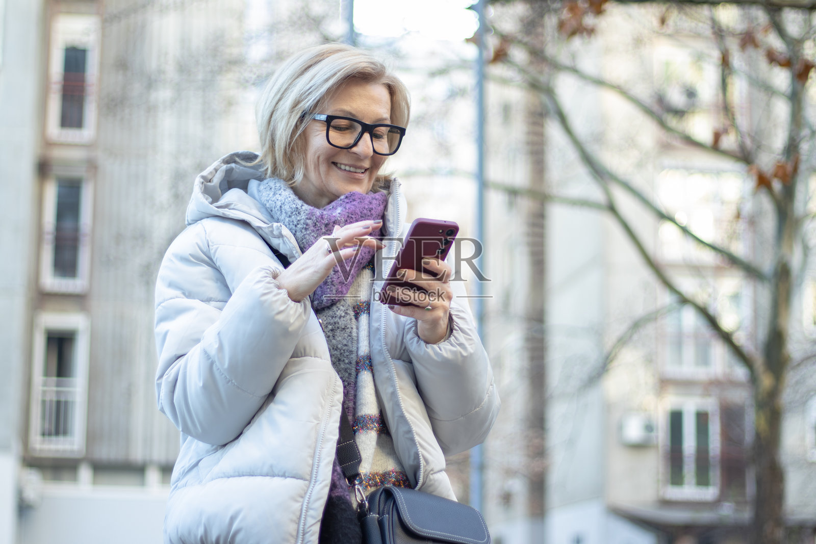 成熟的金发女性戴着眼镜，在公寓楼的公园里走动，冬天的日子里用手机上网。照片摄影图片