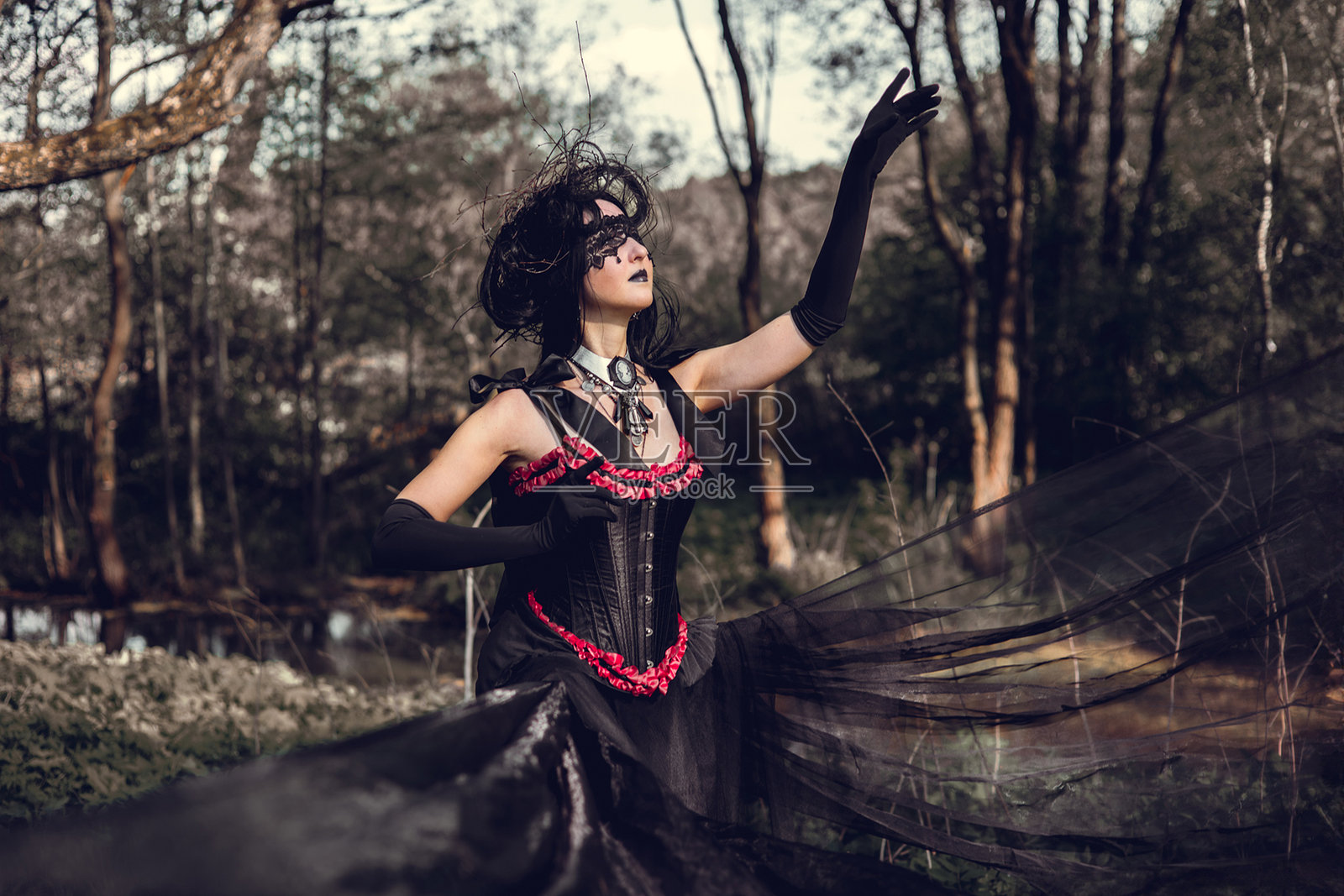 一位年轻女性，形象为女巫和魔法师，站在黑暗的森林中，穿着一条带拖尾的黑色裙子，头上戴着一个头饰。照片摄影图片