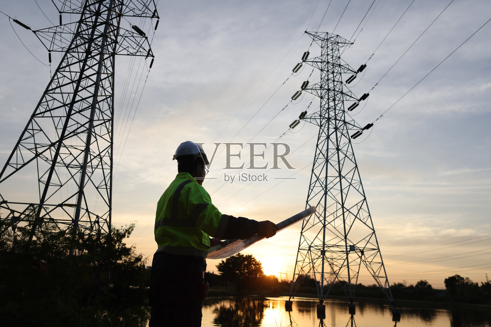 穿着个人防护装备的高级电气工程师在电厂的高压电杆旁绘图，背景是日落。照片摄影图片