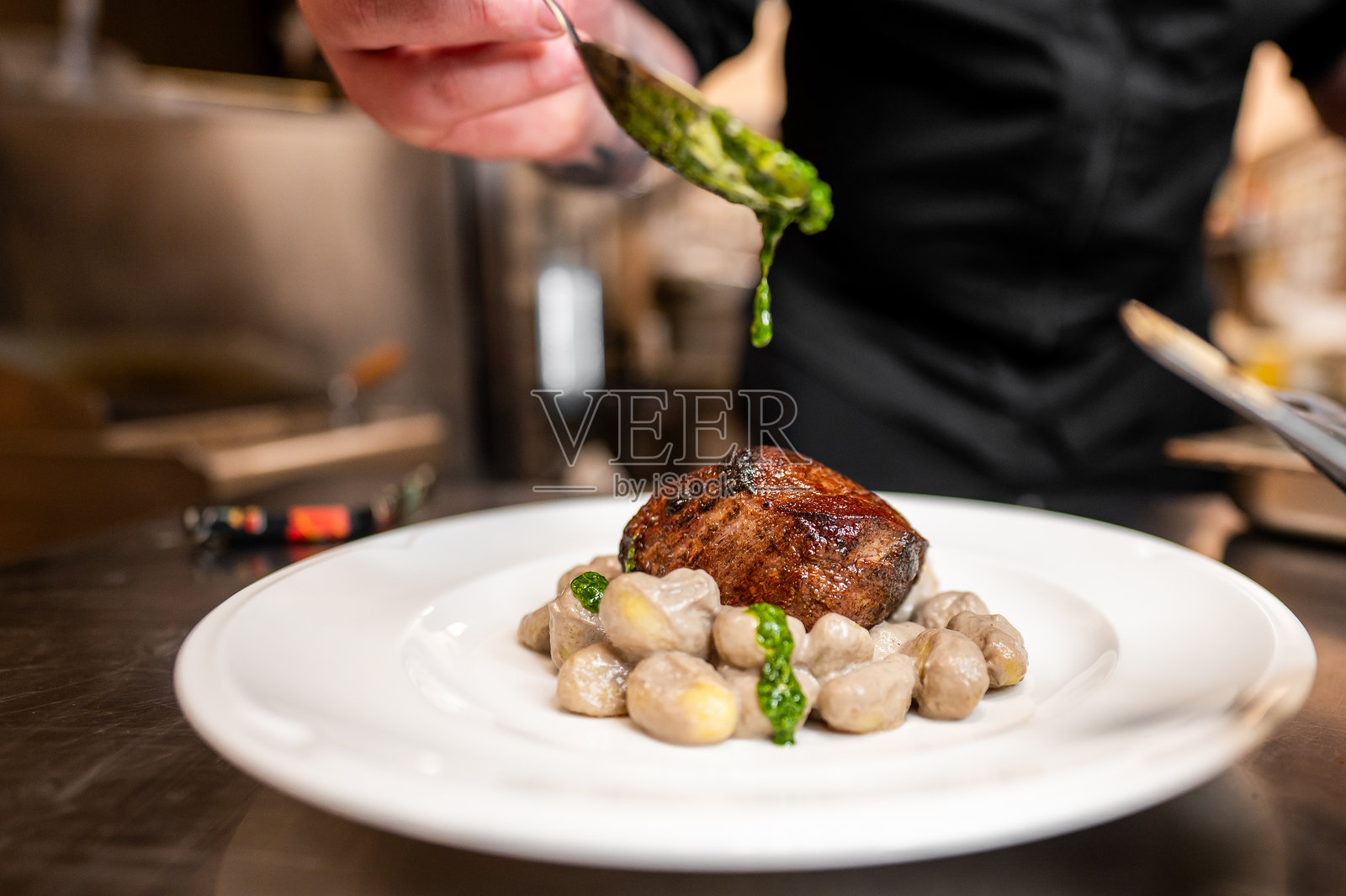 厨师的手小心翼翼地将绿色酱汁淋在精美摆盘的牛排和蘑菇上。这道菜展示了专业厨房环境中的烹饪艺术和对细节的关注。照片摄影图片