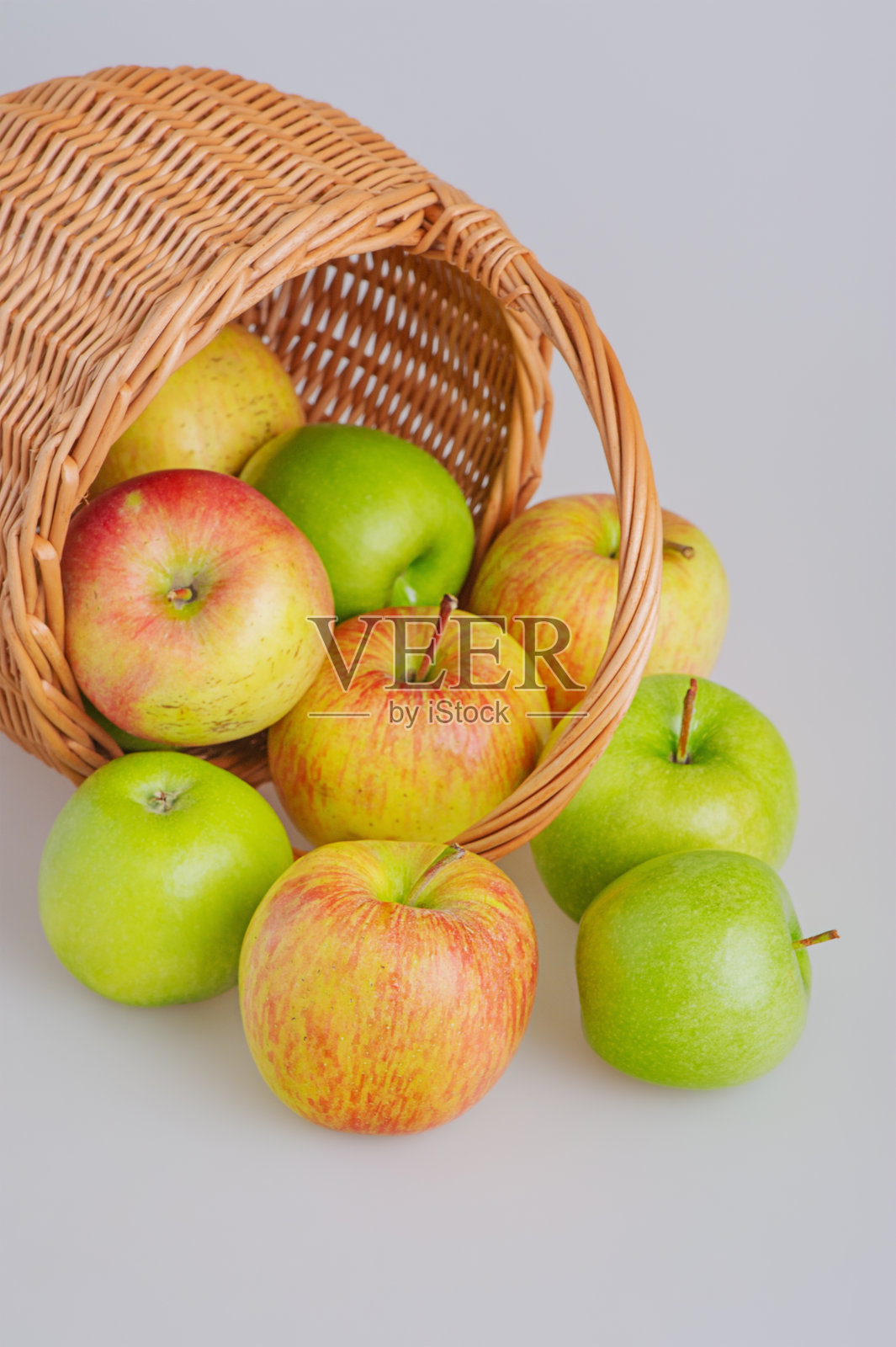 红色和绿色的苹果从一个柳条篮子里洒落在灰色背景上，焦点模糊。水果，苹果照片摄影图片