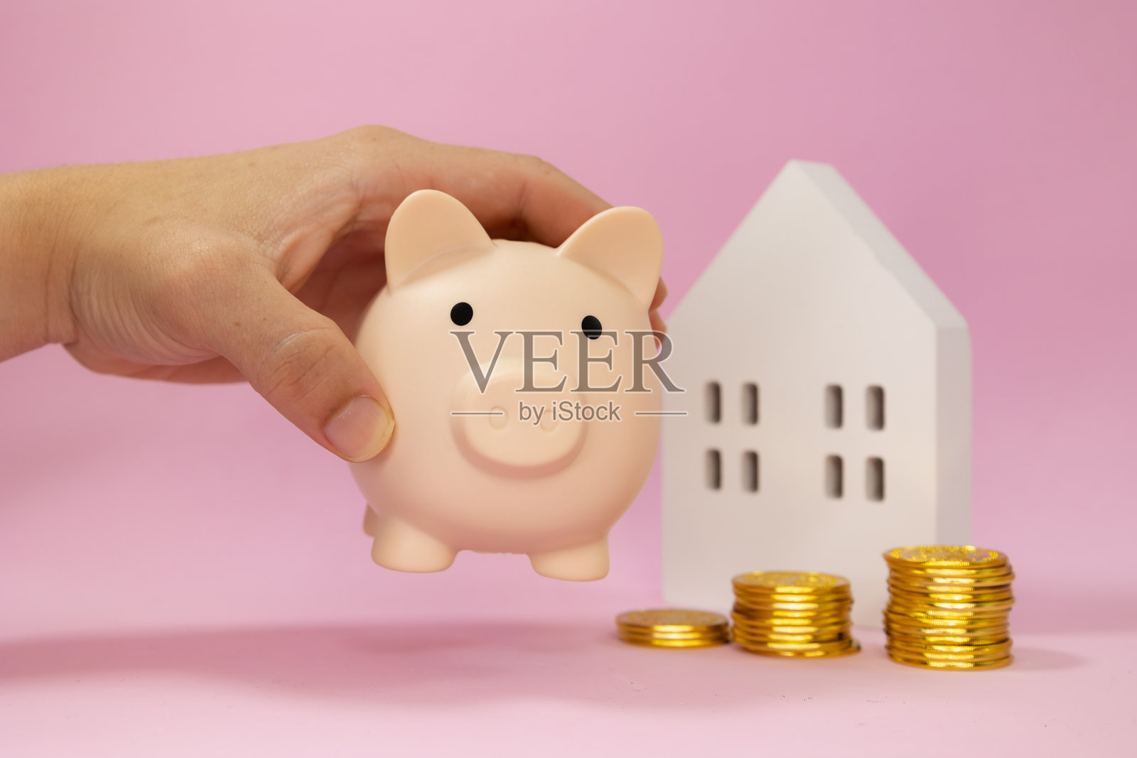 男性手握着一个储蓄罐，周围堆放着金色硬币，旁边有一座白色木屋，背景为粉色。照片摄影图片