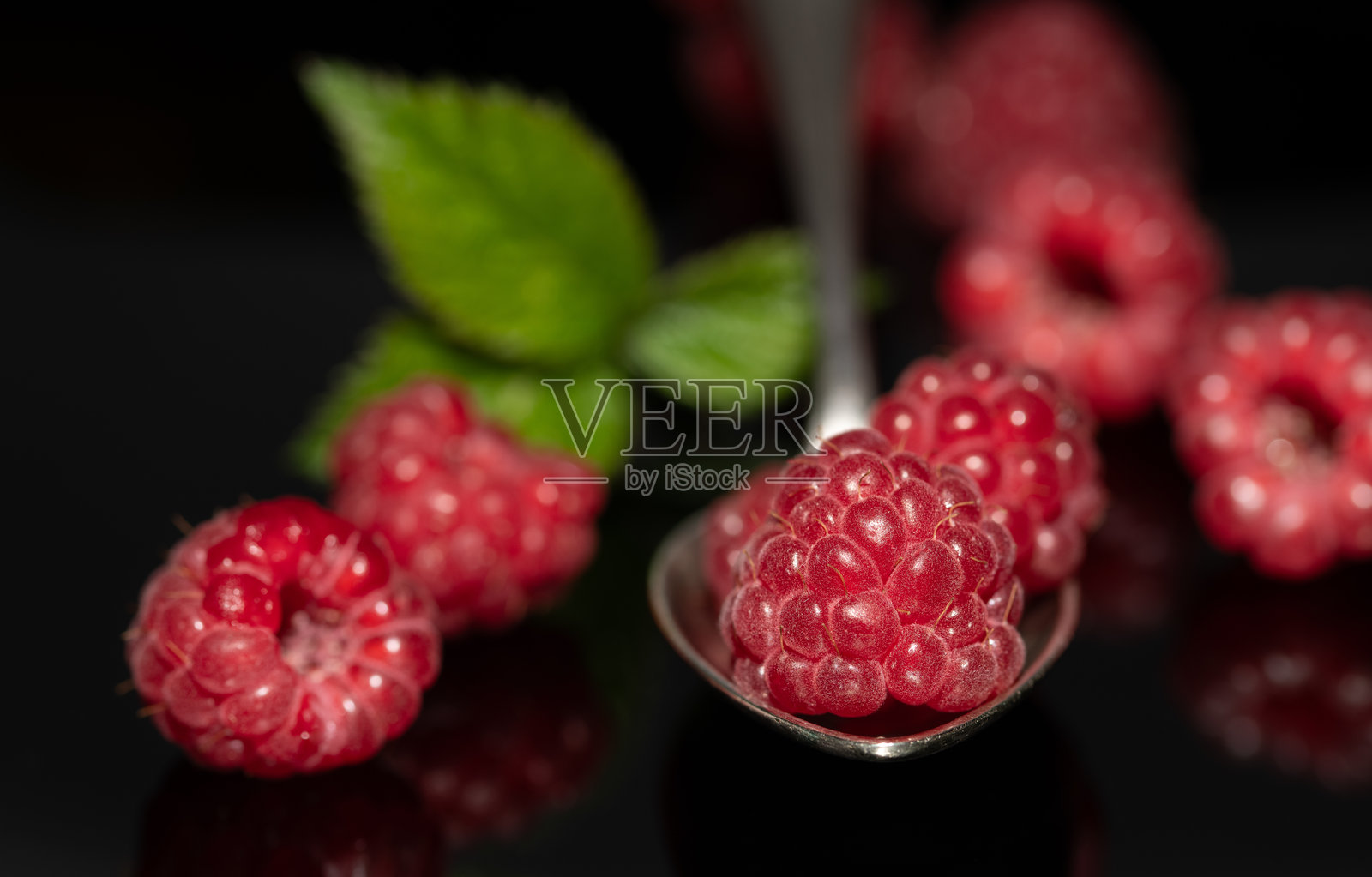 新鲜红色覆盆子的特写。水果放在黑色背景上，形成反射。一颗覆盆子放在金属勺子上。照片摄影图片