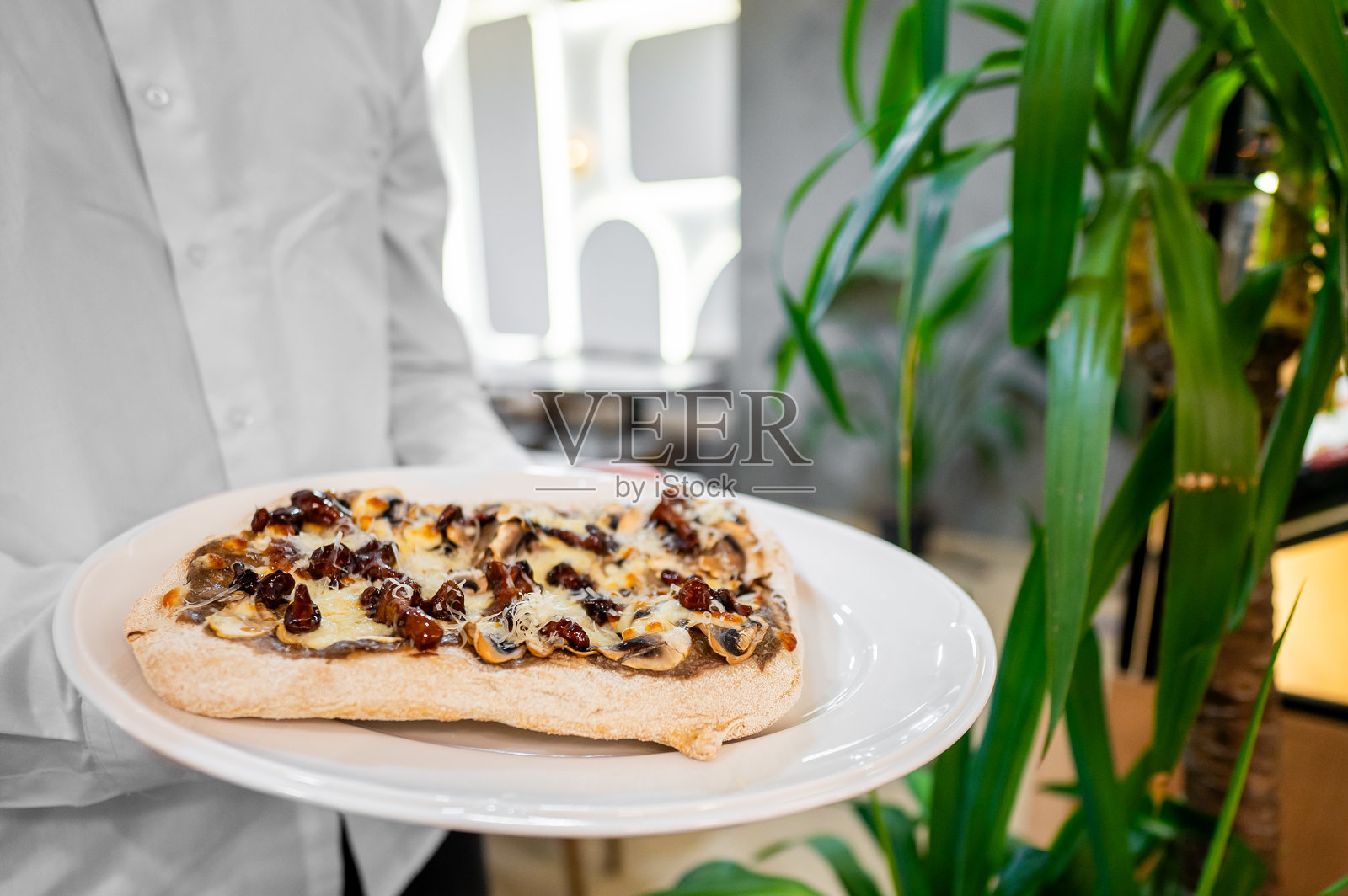 一位服务员手持白色盘子，盘中盛放着美味的焦糖洋葱和奶酪扁面包，背景是时尚的餐厅环境，绿植环绕。照片摄影图片