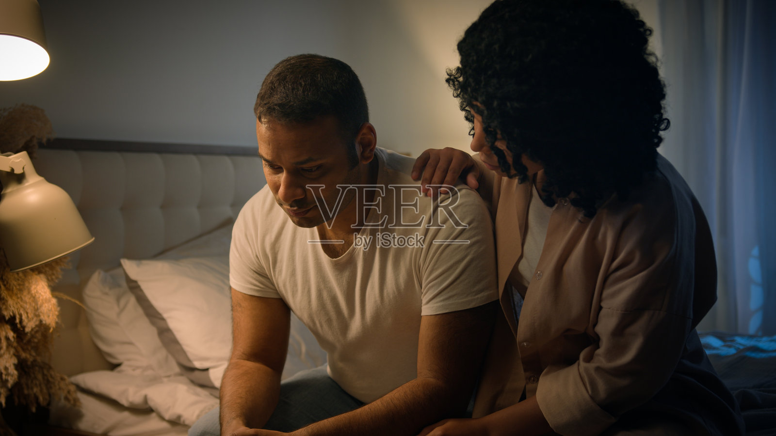 非裔美国人悲伤的男人坐在家中的床上，夜晚感到忧虑和崩溃，因勃起功能障碍而感到后悔，深爱的妻子拥抱他，给予安慰和支持，展现出同情心，面对健康危机。照片摄影图片