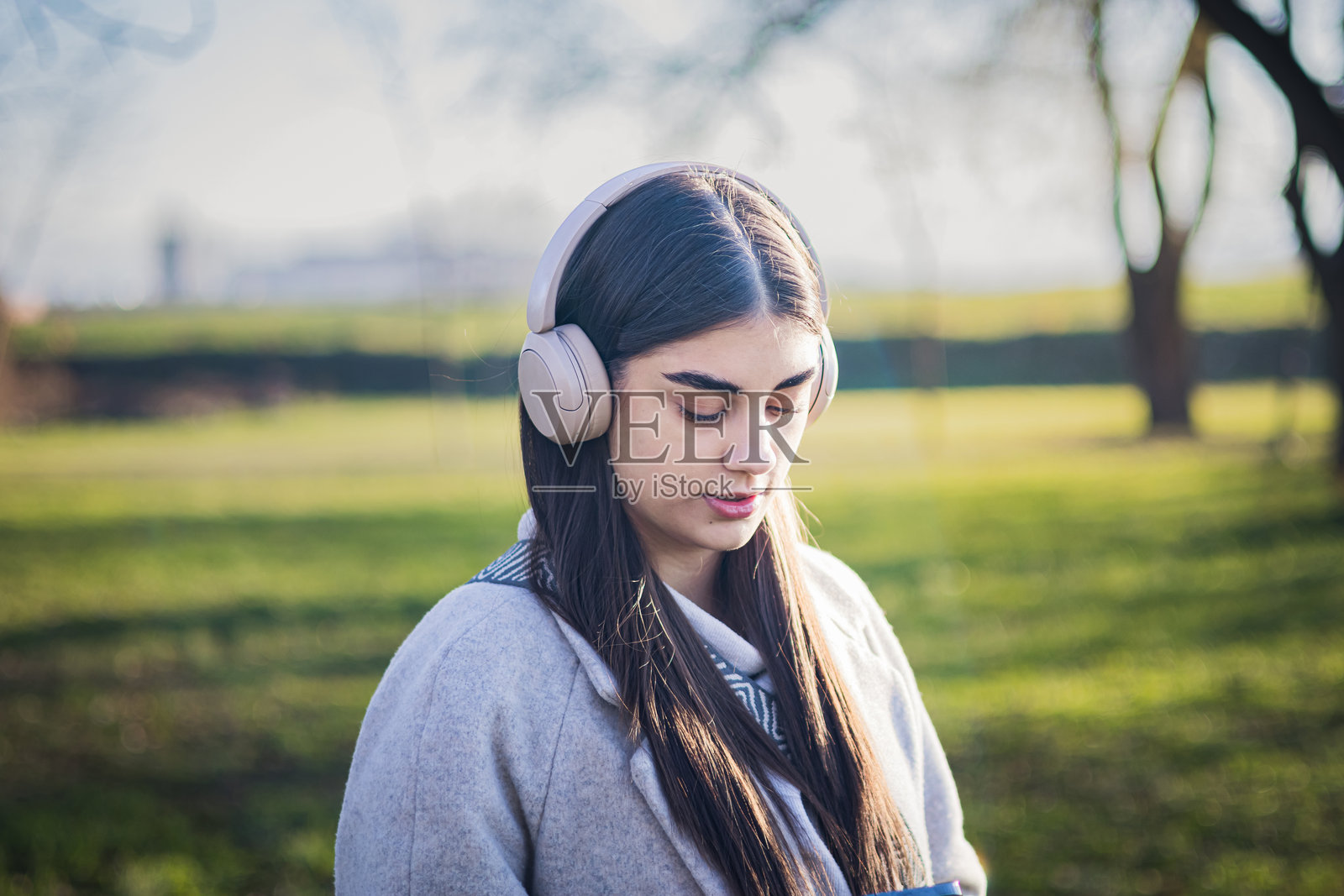 美丽而伤感的年轻女孩戴着耳机，手握手机，在寒冷的冬日公园里独自走着，等待男朋友发来短信。照片摄影图片