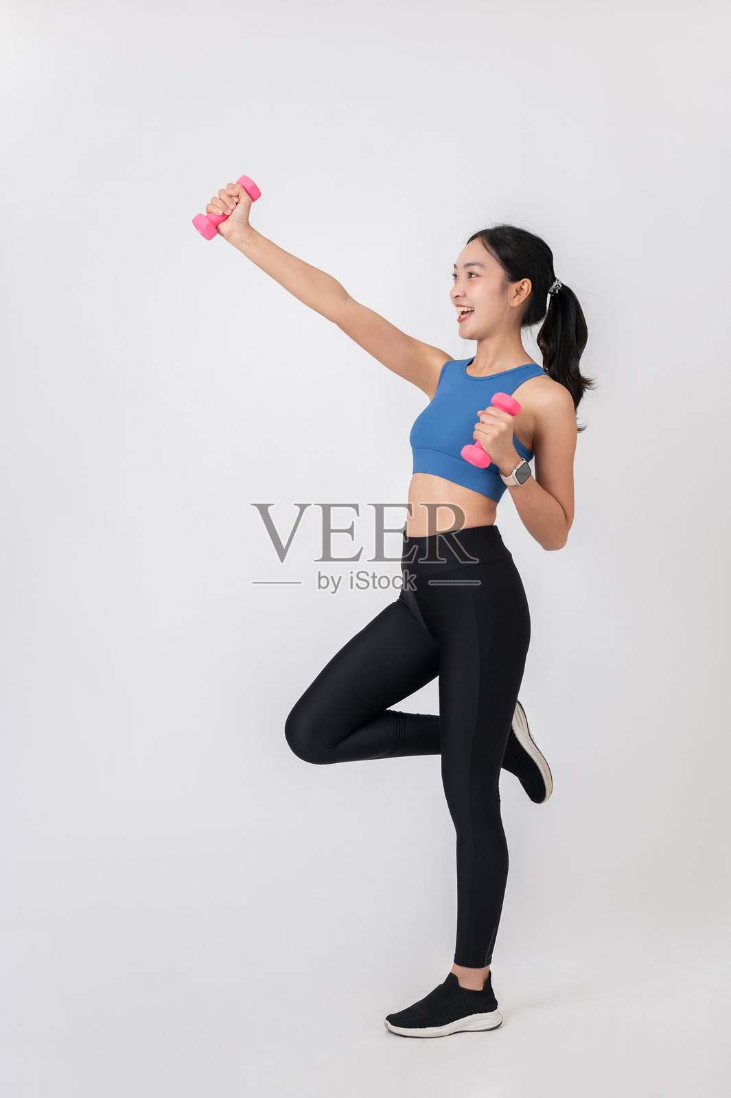一位穿着运动服的年轻亚洲女性在白色摄影棚背景下开心地举着粉色哑铃。照片摄影图片