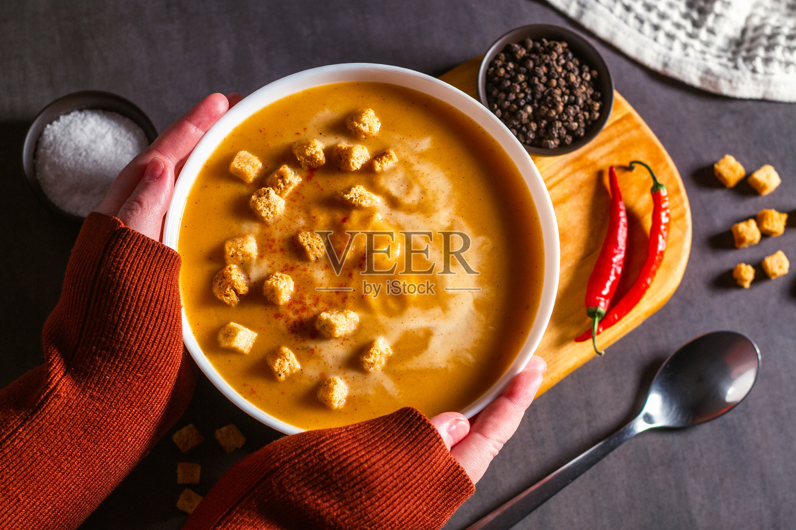 女人手持一碗南瓜汤，旁边有饼干。桌上摆放着奶油汤、辣椒和餐巾。蔬菜汤俯视图。照片摄影图片