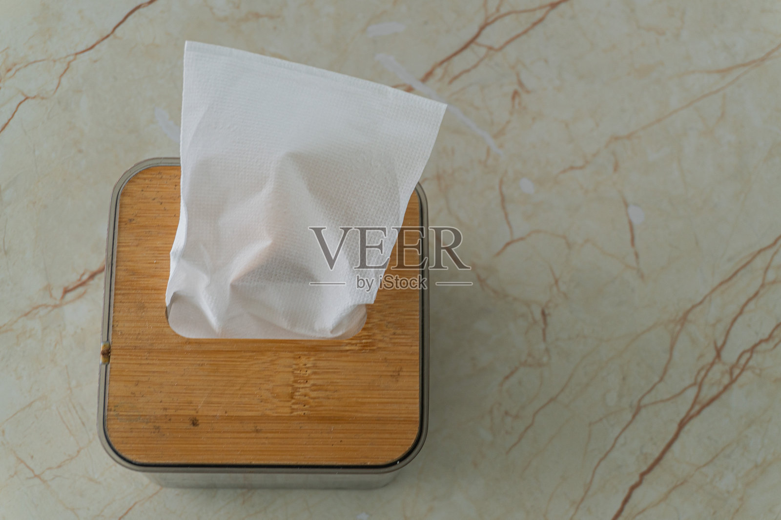 方形纸巾盒配有竹制盖子，放置在浅米色大理石表面上。纸巾盒中抽出一张纸巾。照片摄影图片