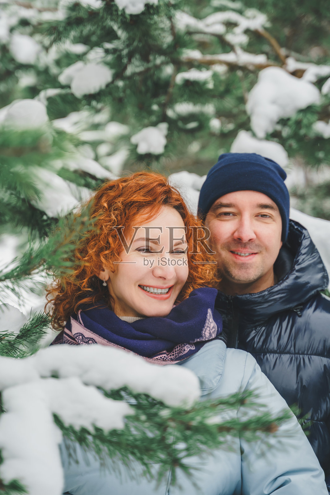 浪漫爱情故事。快乐的中年千禧夫妇，冬季雪地公园里一位红发女子。浪漫约会，亲吻，拥抱。散步，享受乐趣。时尚的蓝色衣服，夹克，帽子。照片摄影图片