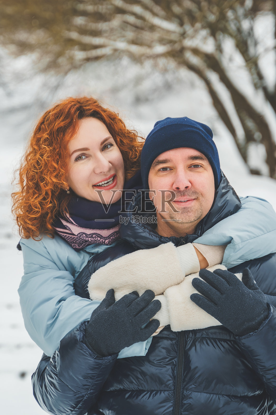 浪漫爱情故事。快乐的中年千禧夫妇，冬季雪地公园里一位红发女子。浪漫约会，亲吻，拥抱。散步，享受乐趣。时尚的蓝色衣服，夹克，帽子。照片摄影图片