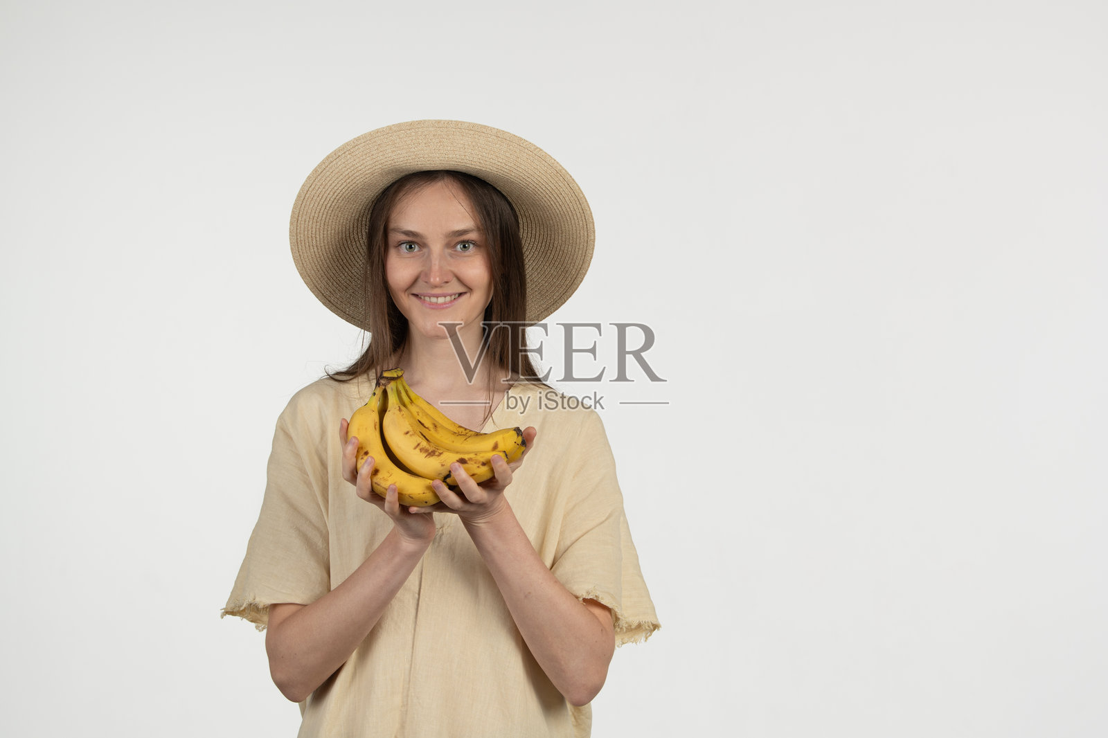 一位长发女性微笑着，手中拿着一束成熟的香蕉。她穿着浅色上衣，戴着宽边草帽，背景为中性色调。照片摄影图片
