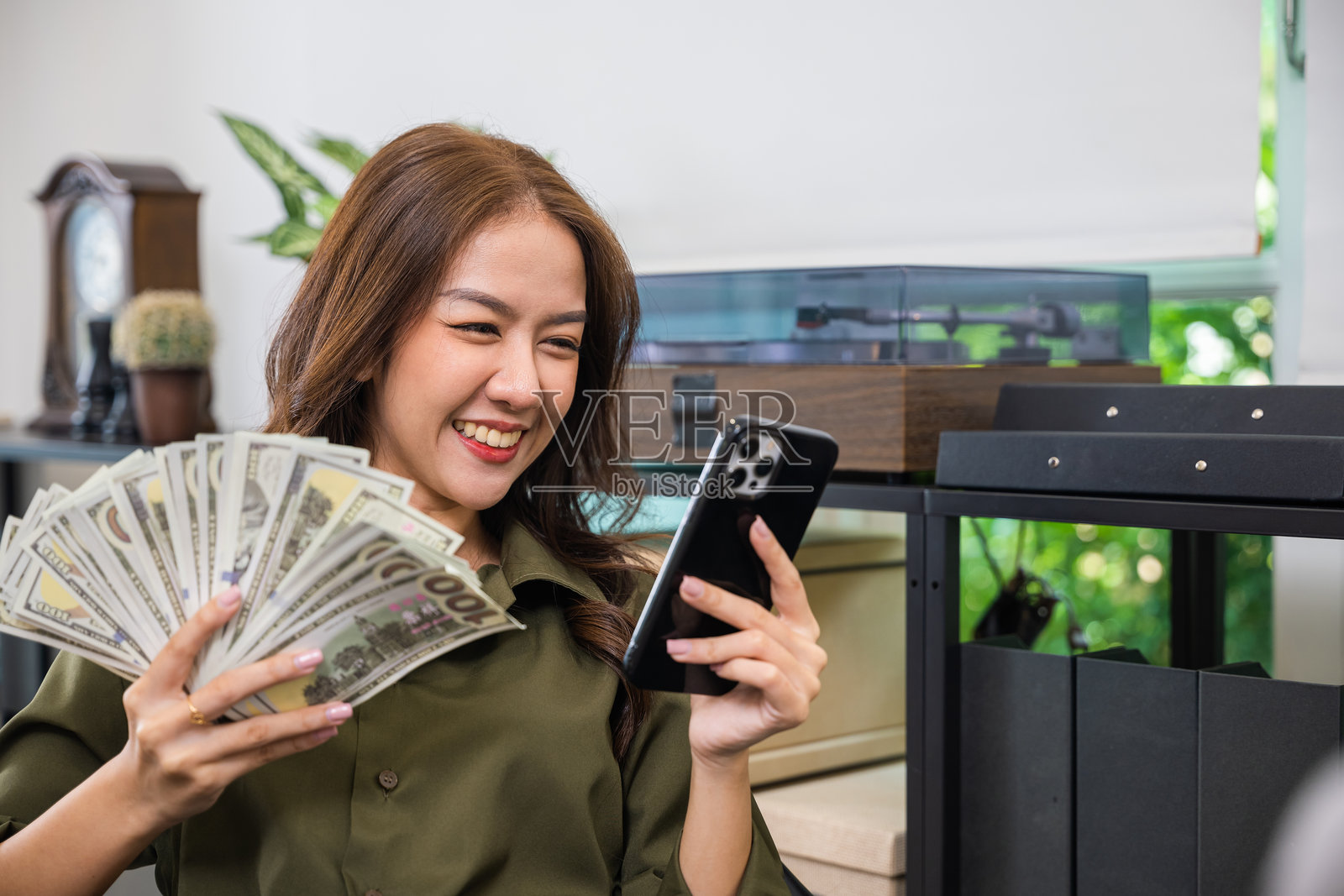 一位微笑的亚洲女性员工，手持美元钞票和手机，坐在家中的办公桌前。照片摄影图片