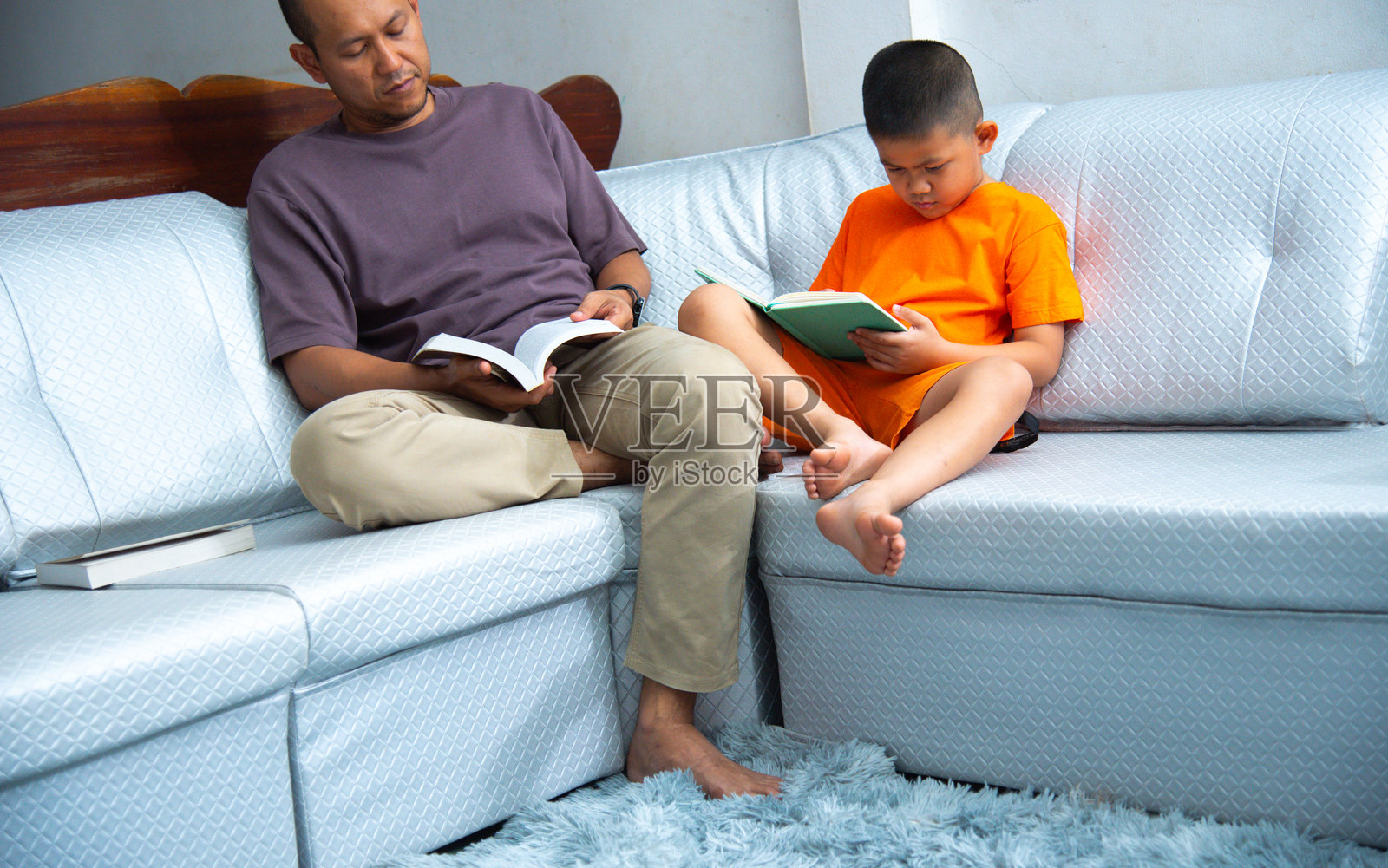 父亲和儿子坐在舒适的沙发上，一起合作完成作业。男孩手里拿着一本书，而父亲则用教科书来辅助他们的学习。教育，家庭概念照片摄影图片