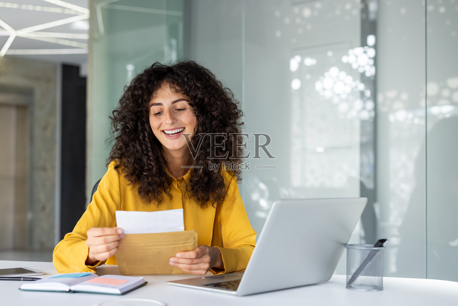 微笑的拉丁美洲女商人在现代办公室的桌子上阅读信件，桌上有笔记本电脑和咖啡。专注的专业人士享受着积极的消息或成功的电子邮件沟通。照片摄影图片