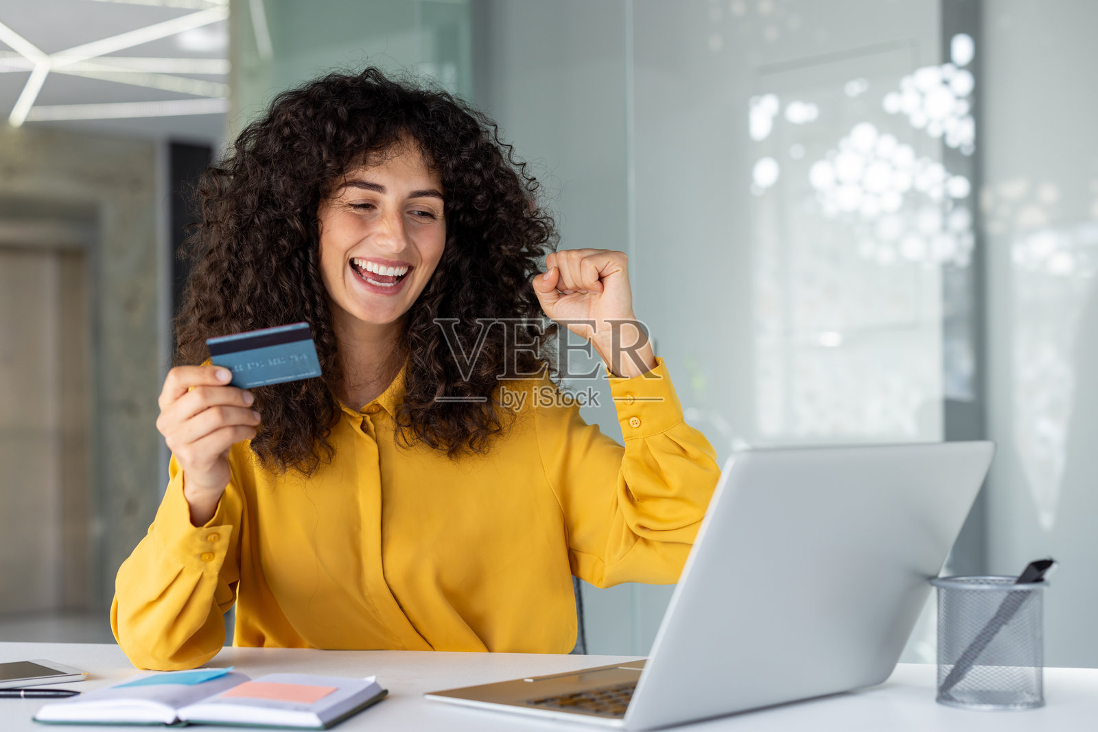 穿着黄色衬衫的快乐女性在工作桌前庆祝成功，手持信用卡并使用笔记本电脑，展示在线购物、财务自由和快乐成就的概念。照片摄影图片