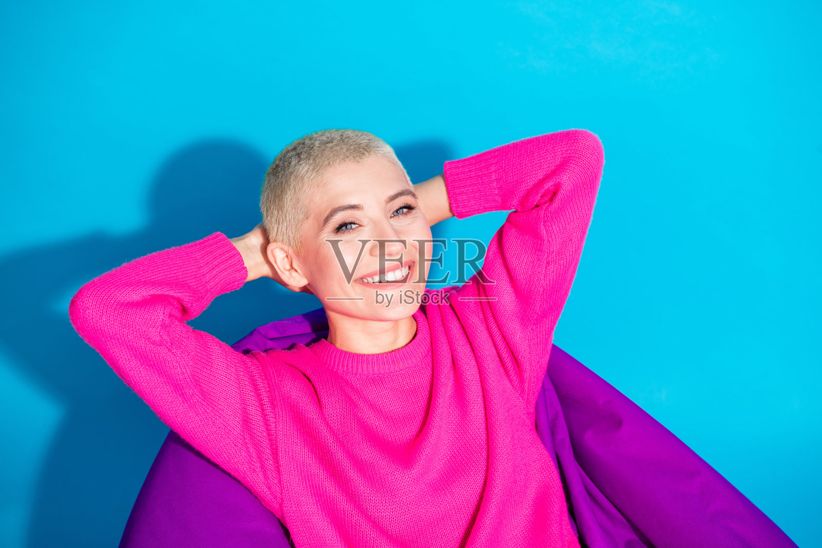 微笑的年轻短发女性穿着粉色毛衣，靠在鲜艳的蓝色背景上放松。照片摄影图片