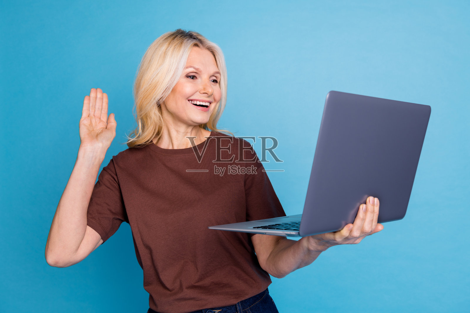 可爱的老年女性穿着时尚的棕色衣服，使用现代设备，背景为蓝色。照片摄影图片