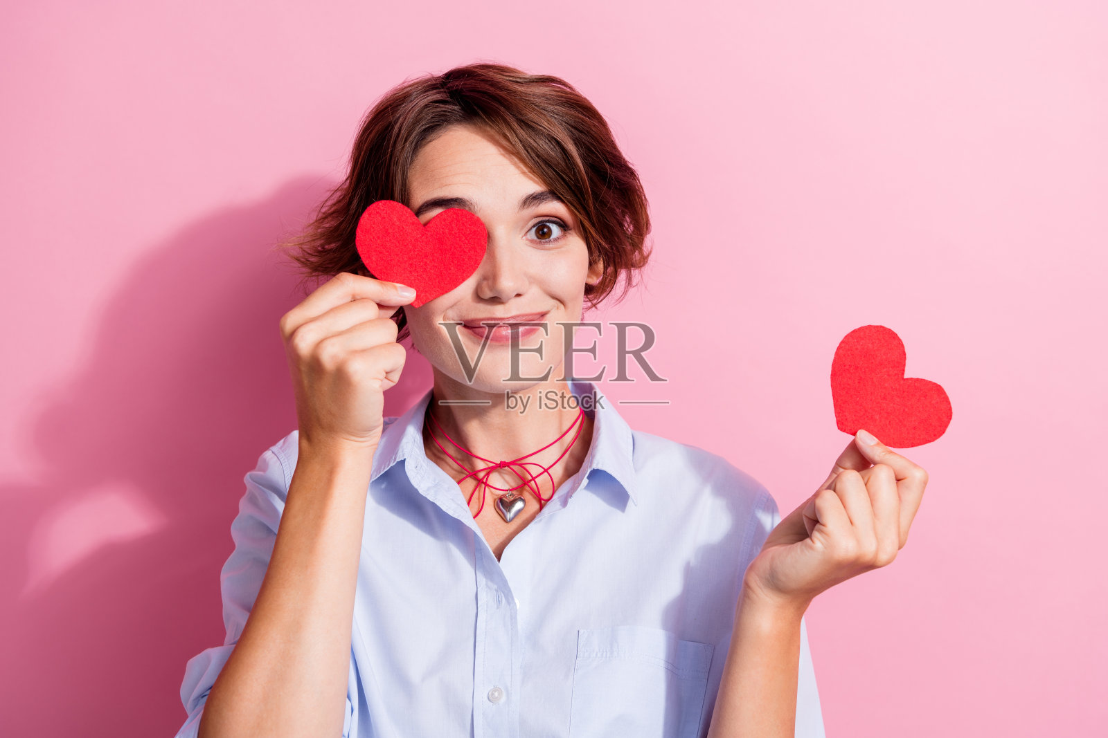漂亮积极的女孩用小红纸心遮住一只眼睛，背景为粉色。照片摄影图片