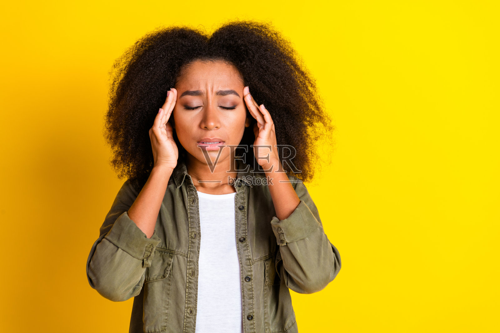 一位疲惫痛苦的女孩，留着卷发，穿着卡其色衬衫，双手放在头上，闭着眼睛，背景为黄色。照片摄影图片