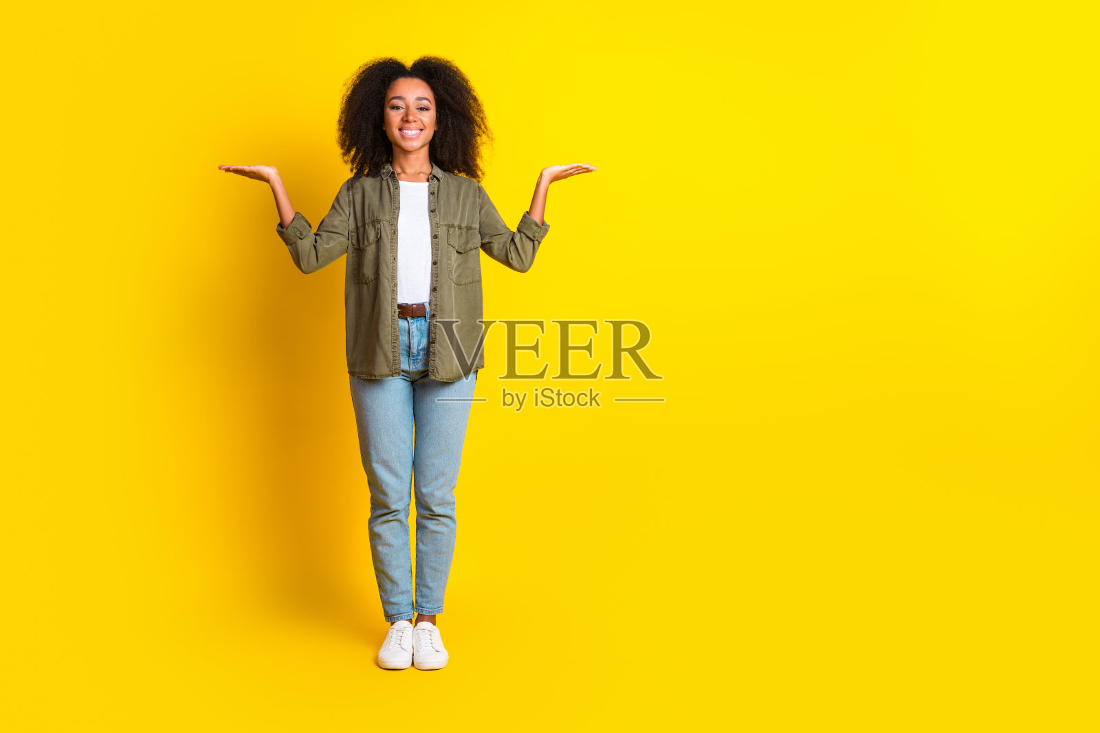 全身照，愉快的女孩穿着卡其色衬衫和牛仔裤，双手比较产品，背景为黄色，空白区域。照片摄影图片