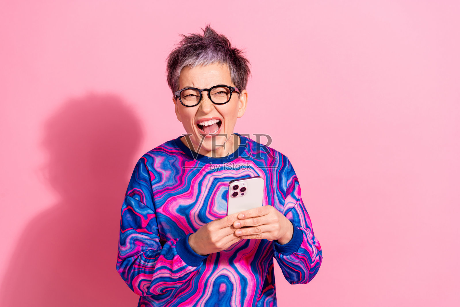 可爱的退休女士在粉色背景下兴奋地尖叫，手持设备，穿着蓝色多彩的服装。照片摄影图片