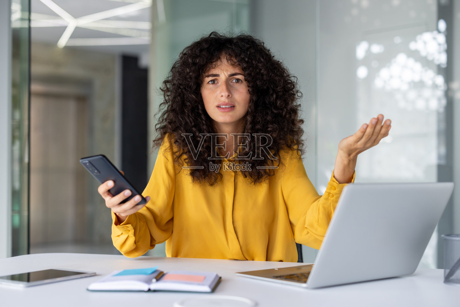 沮丧的卷发女商人在现代办公室的桌子前坐着，穿着黄色衬衫，手持智能手机，困惑地做着手势，同时使用笔记本电脑。照片摄影图片