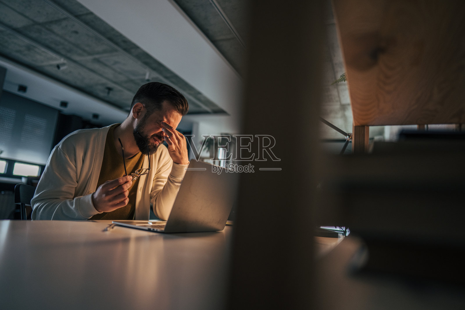疲惫且过度劳累的男子坐在昏暗的家庭办公室里，面前摆着一台笔记本电脑。照片摄影图片