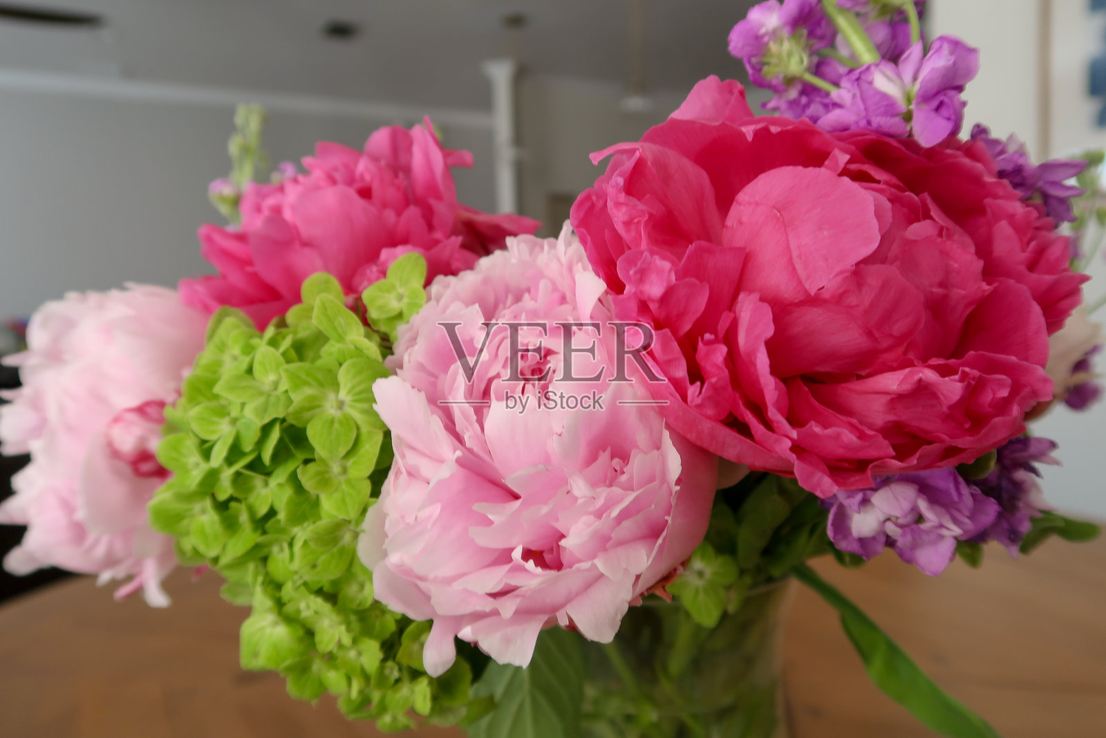 一个生动的花卉布置，包含鲜艳的粉色牡丹、柔和的粉色花朵、绿色绣球花和紫色点缀，放置在一个柔和灯光的室内环境中的花瓶里。照片摄影图片