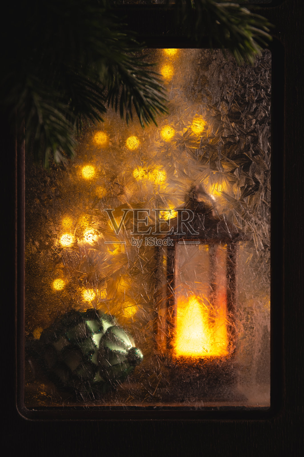 在冰冻的窗户后，一盏发出温暖光芒的圣诞灯笼，装饰着小彩灯、松枝和一个装饰球，营造出温馨的节日氛围。照片摄影图片