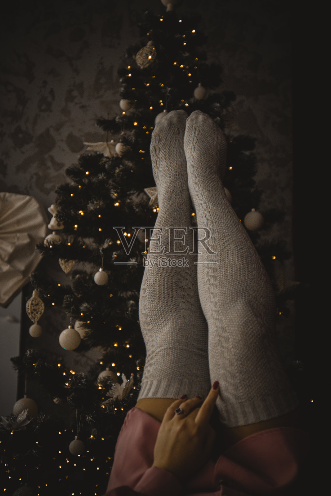 温馨的冬季氛围：一位穿着温暖针织袜子的女性躺在圣诞树前，灯光闪烁，洋溢着节日的气氛和家的温暖。照片摄影图片