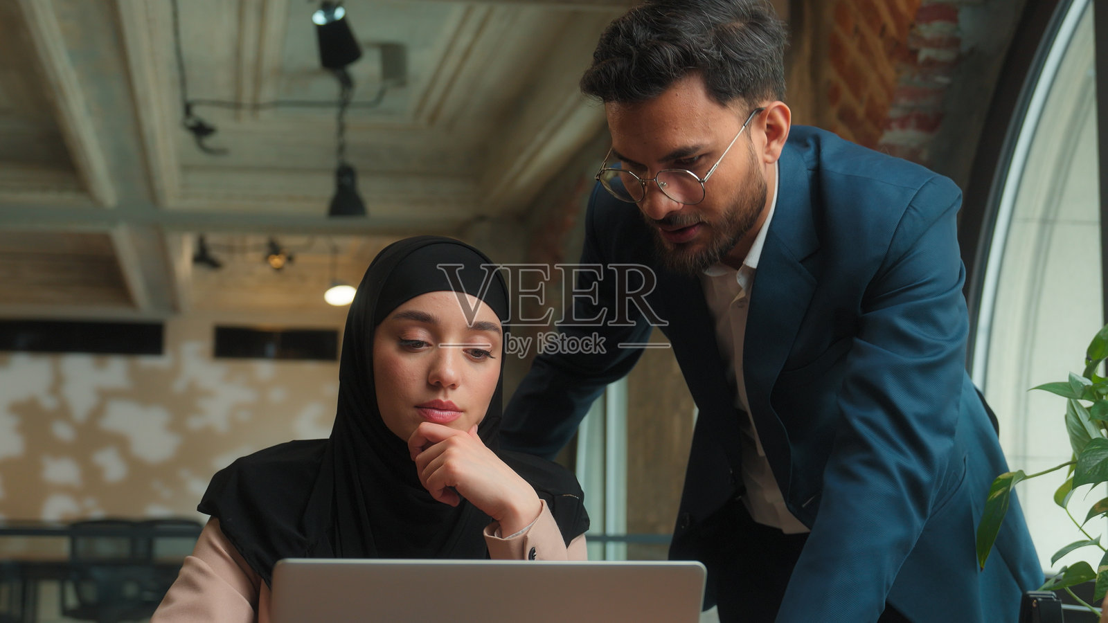 男性商人教导女性穆斯林员工在办公室佩戴头巾，导师领导帮助她使用笔记本电脑，咨询支持学生，教授计算机任务，阿拉伯男士与女性同事团队合作。照片摄影图片