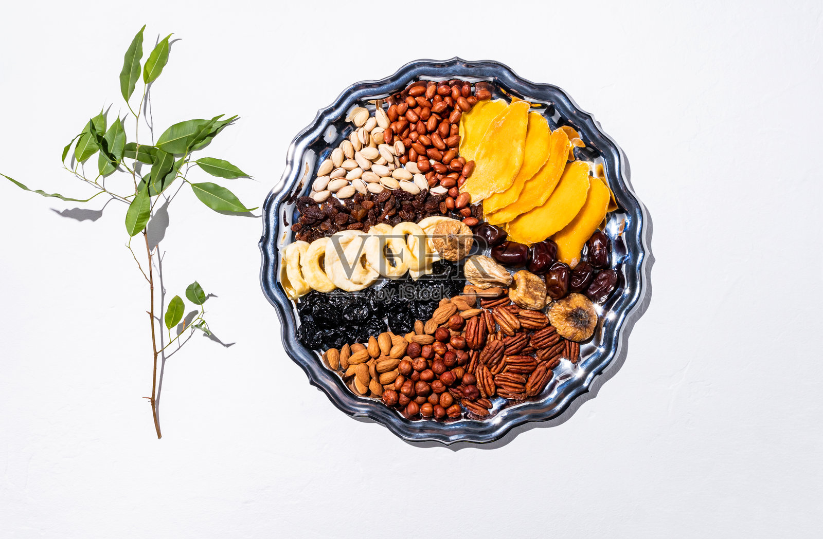 一只美丽的银盘，上面摆放着各种干果和坚果，背景为白色，旁边有一根绿色树枝。俯视图。图比什瓦特的概念。照片摄影图片