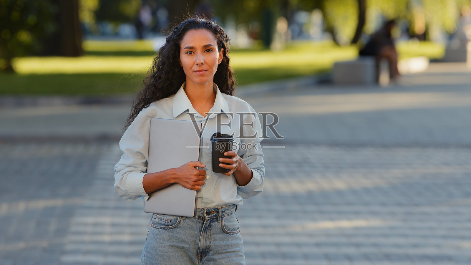 年轻美丽成功的棕发女孩，卷发的女商人，手持笔记本电脑和咖啡，微笑着站在斑马线前，背景是交通，面向镜头，喝着茶，摆姿势在街上。照片摄影图片