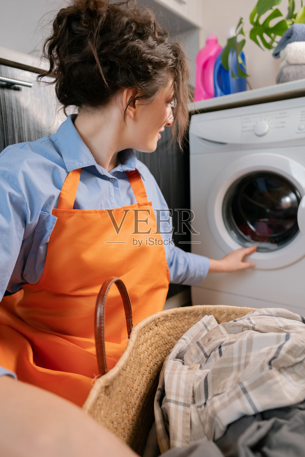 穿着橙色围裙的女性启动洗衣机，现代洗衣机具备智能功能和特点。照片摄影图片