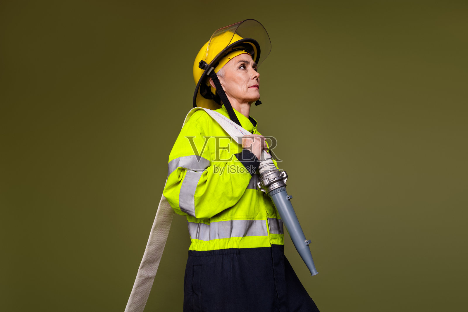 成熟的女性消防员穿着反光制服和头盔，手持水管，背景为卡其色，象征着力量照片摄影图片
