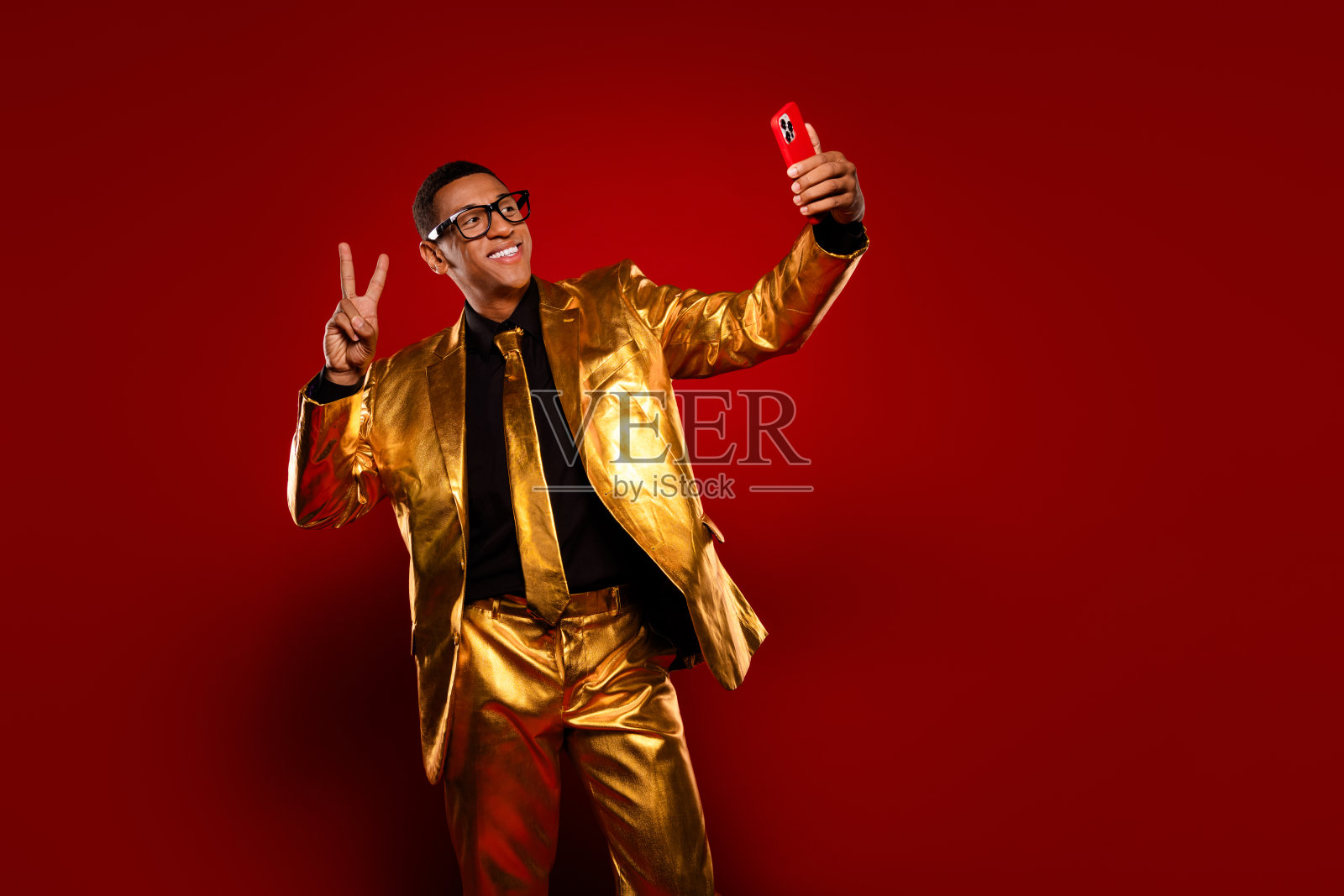 自信的年轻男子穿着金色西装，在鲜艳的红色背景前比出和平手势自拍照片摄影图片