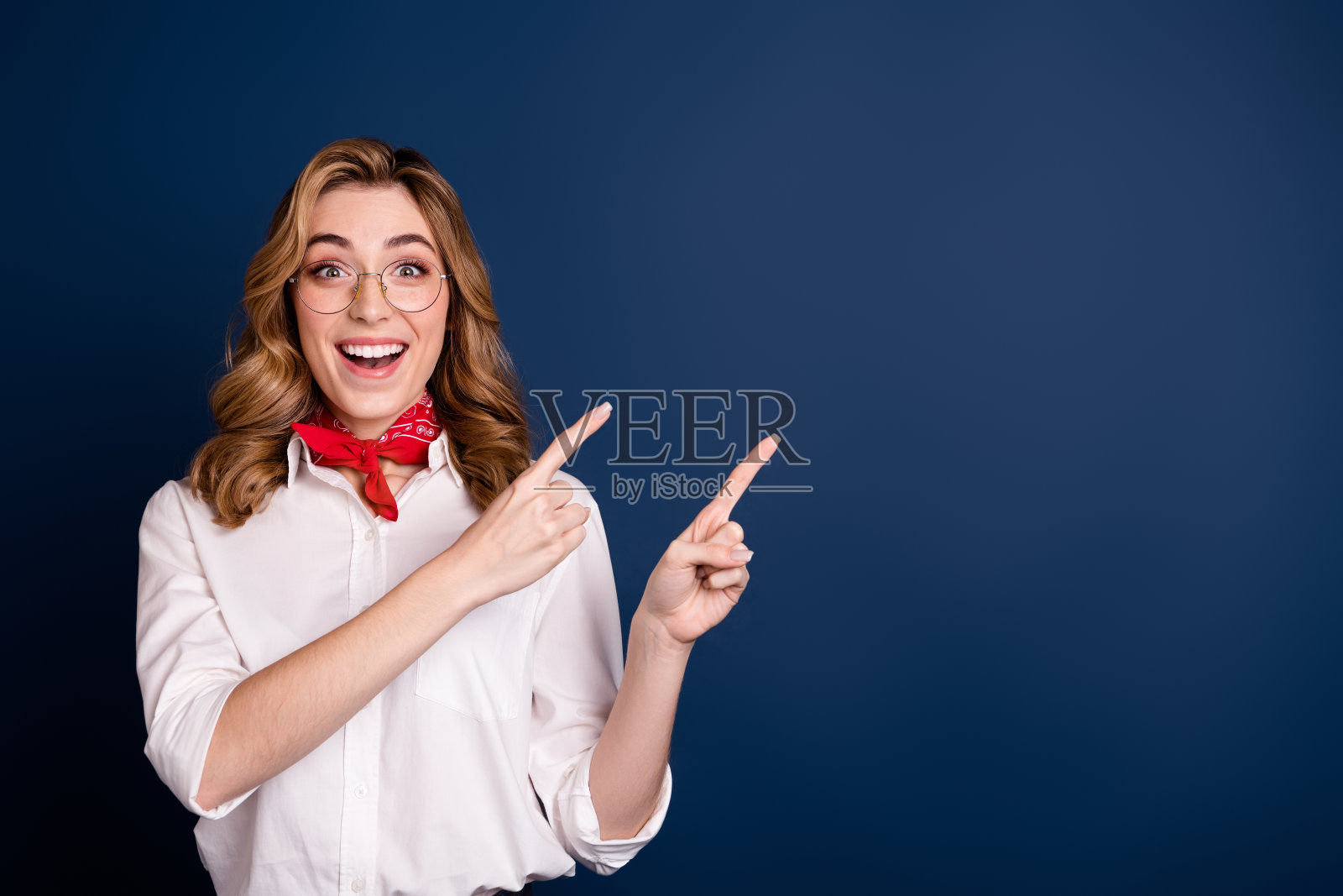 快乐的年轻女性兴奋地指着画外的某个东西，佩戴着时尚的红色围巾和眼镜，背景为深蓝色。照片摄影图片