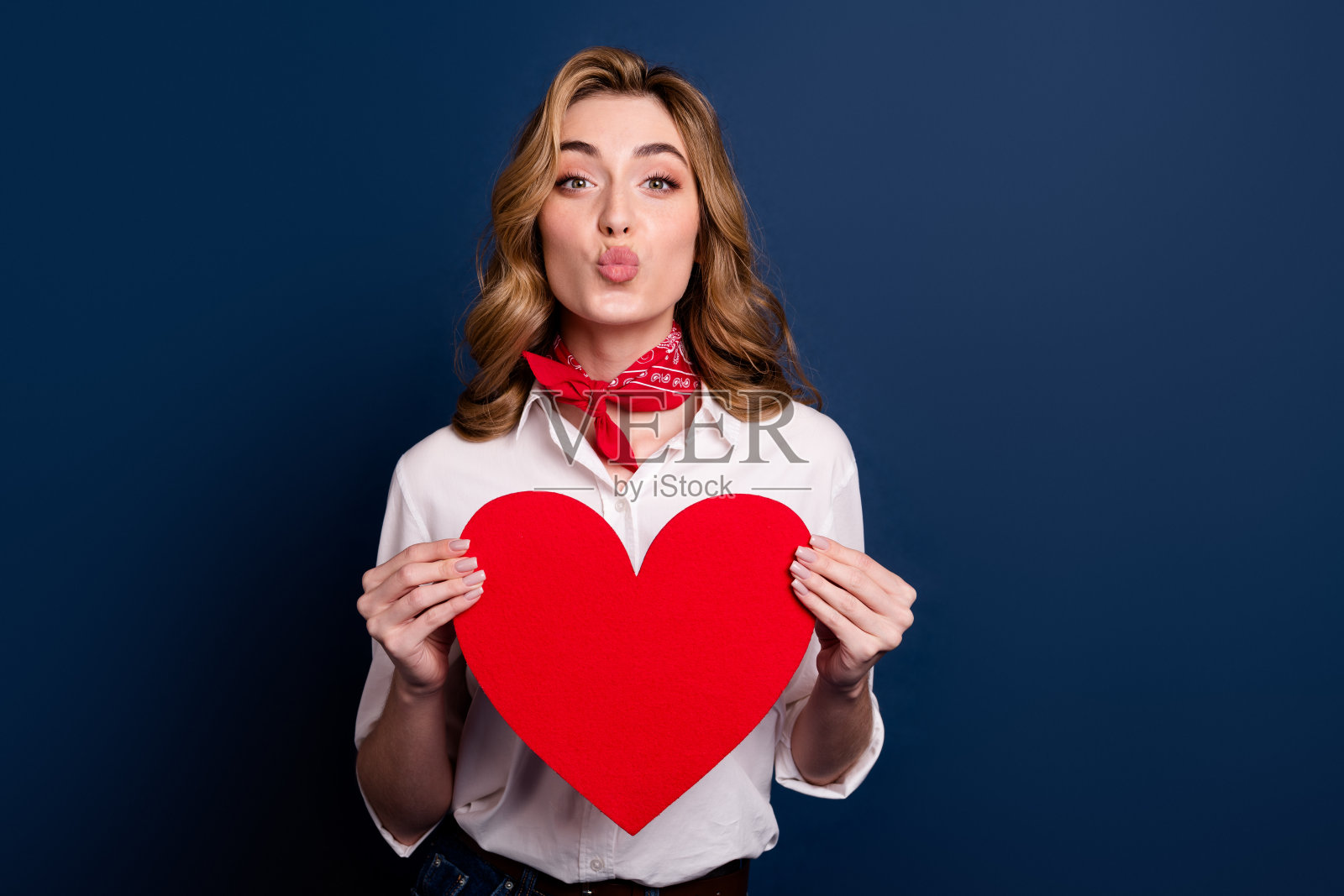 年轻女性在深蓝色背景下微笑，手持心形符号，穿着休闲时尚，系着红色围巾。照片摄影图片