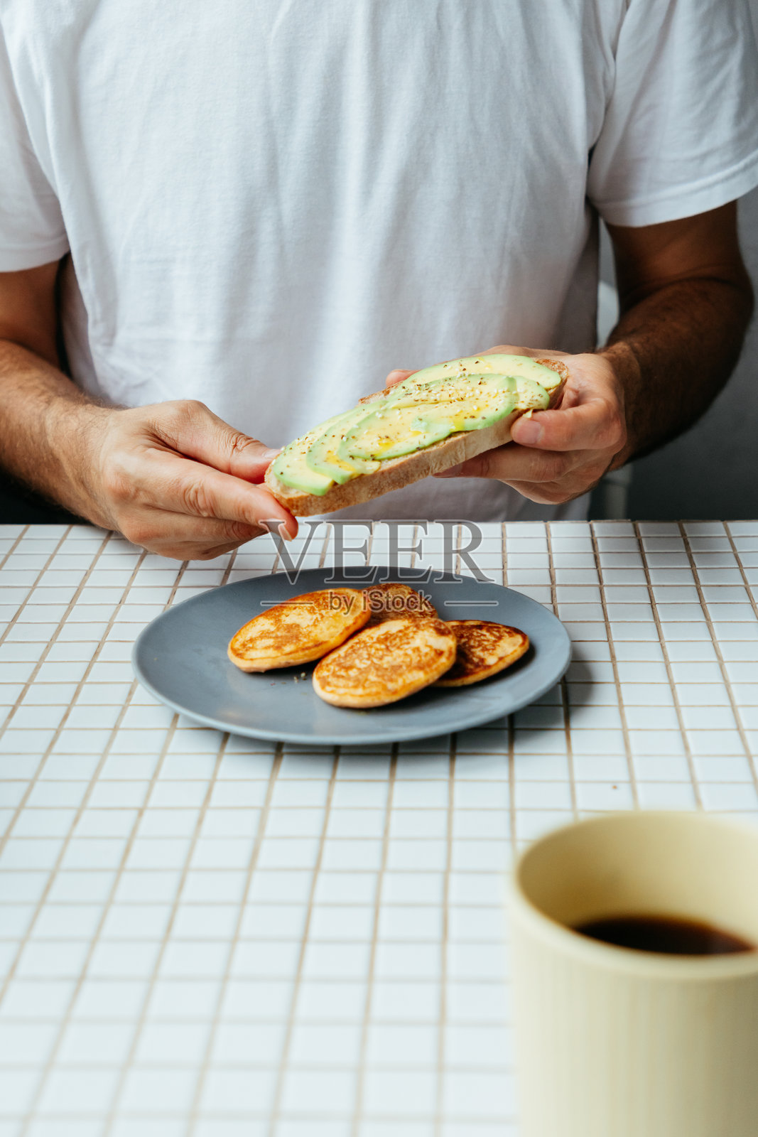 男人在家里的厨房吃早餐。盘子里有鳄梨吐司、煎饼和黑咖啡。照片摄影图片